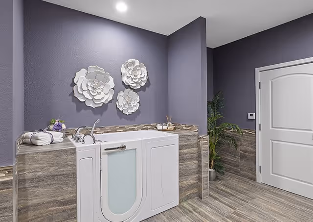 A modern bathroom with a walk-in bathtub featuring a glass door. The walls are painted purple with decorative white flower wall art above the tub. The floor and lower walls are covered with grayish-brown tiles. There is a white door on the right side and a potted plant next to it. Towels and a small flower arrangement are placed on the edge of the bathtub.
