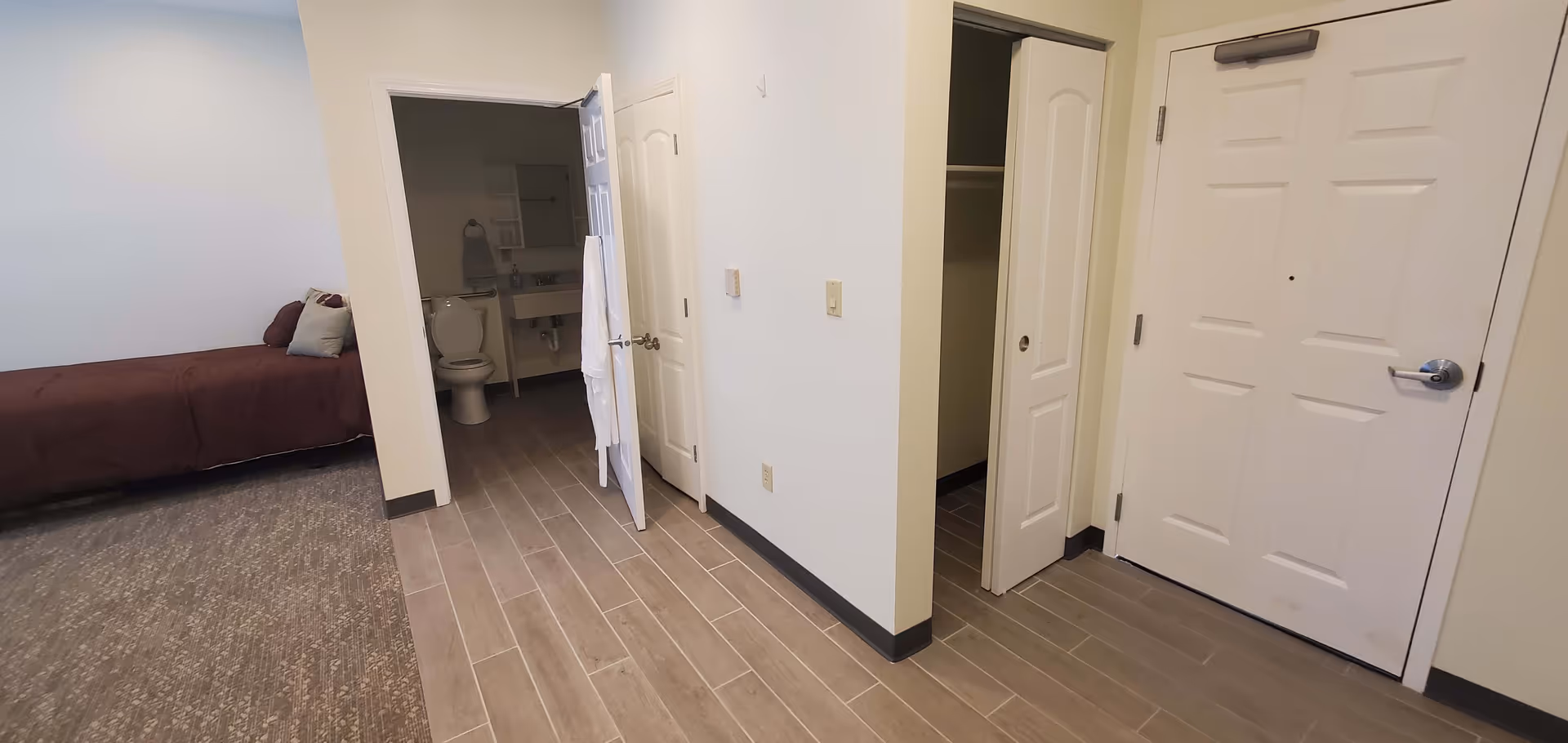 Interior view of a senior living facility room showing a bed with pillows on the left, an open door leading to a bathroom with a toilet and sink, a closet with sliding doors, and a main entrance door on the right. The floor has a combination of carpet and tile.