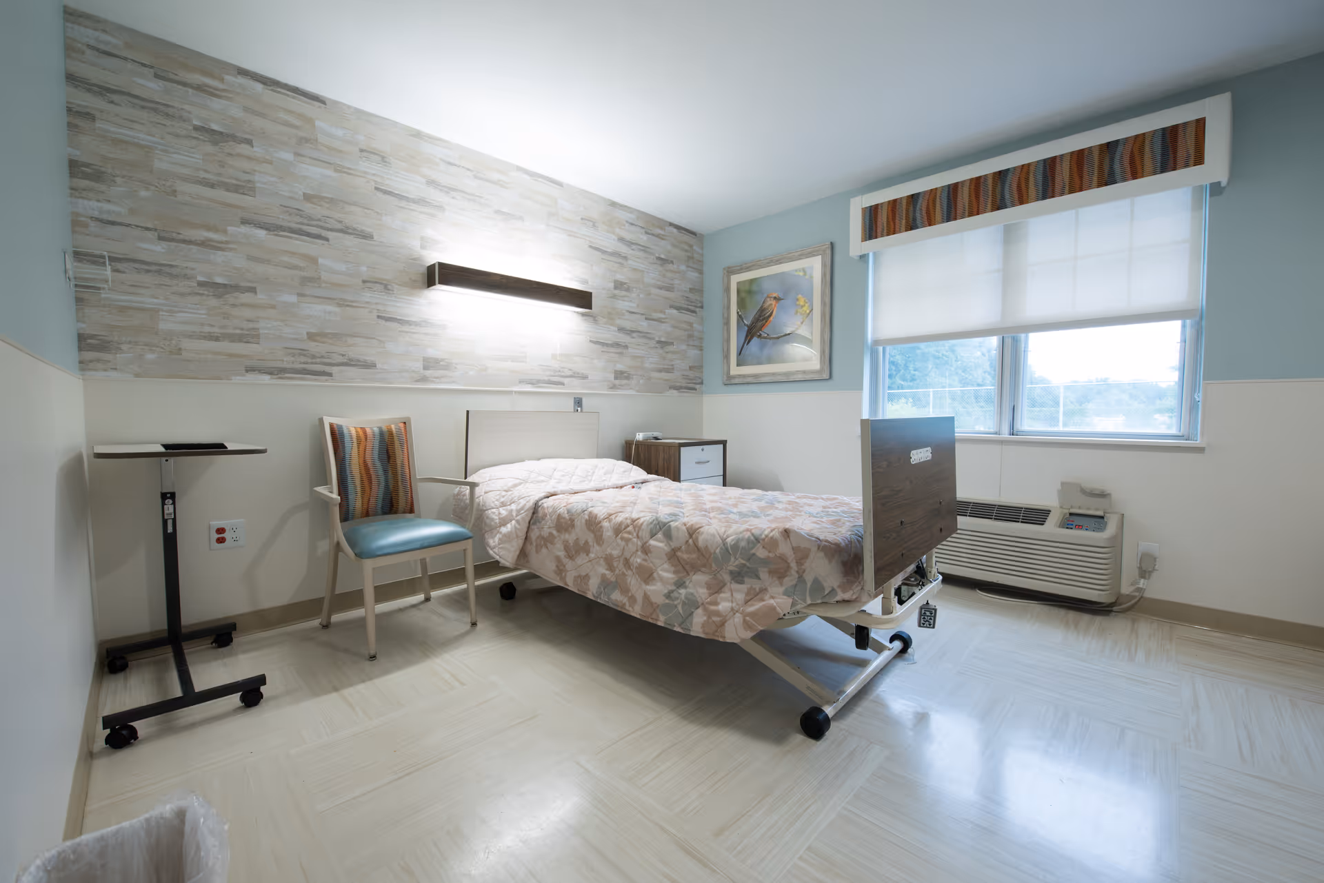 A clean and simple patient room in a healthcare facility featuring a single hospital bed with floral bedding, a striped upholstered chair, a small bedside table, a rolling overbed table, a window with a colorful valance and a white roller shade, and a wall-mounted light above a textured accent wall. There is also an air conditioning unit below the window and a framed picture of a bird on the wall.