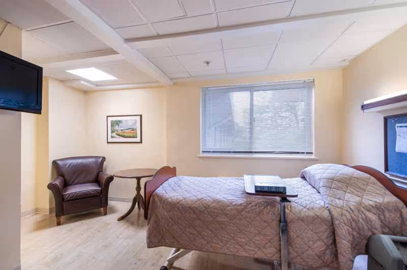 A senior living facility bedroom with a single bed covered in a beige quilt, a small overbed table with a remote control on it, a brown leather armchair, a round wooden side table, a framed picture on the wall, a window with blinds, and a wall-mounted television.