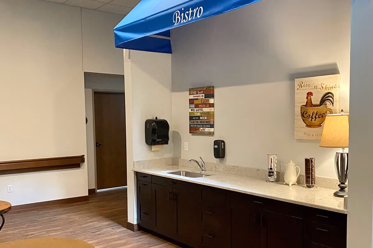 Interior view of a senior living facility's bistro area with a countertop, sink, dark wooden cabinets, a lamp, and decorative wall art including a coffee-themed sign and an inspirational quote. The floor is wooden, and there is a blue awning labeled 'Bistro' above the countertop.
