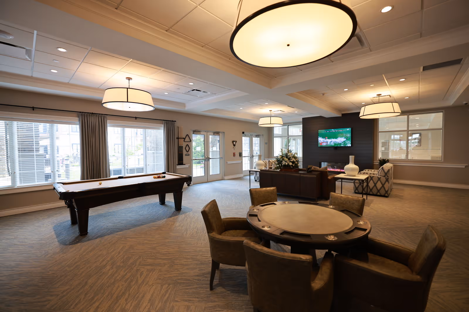 A spacious senior living common area featuring a pool table, a round card table with four chairs, a seating area with a sofa and armchairs around a modern fireplace, and a wall-mounted TV. Large windows and glass doors allow natural light to fill the room, which is illuminated by multiple ceiling lights.