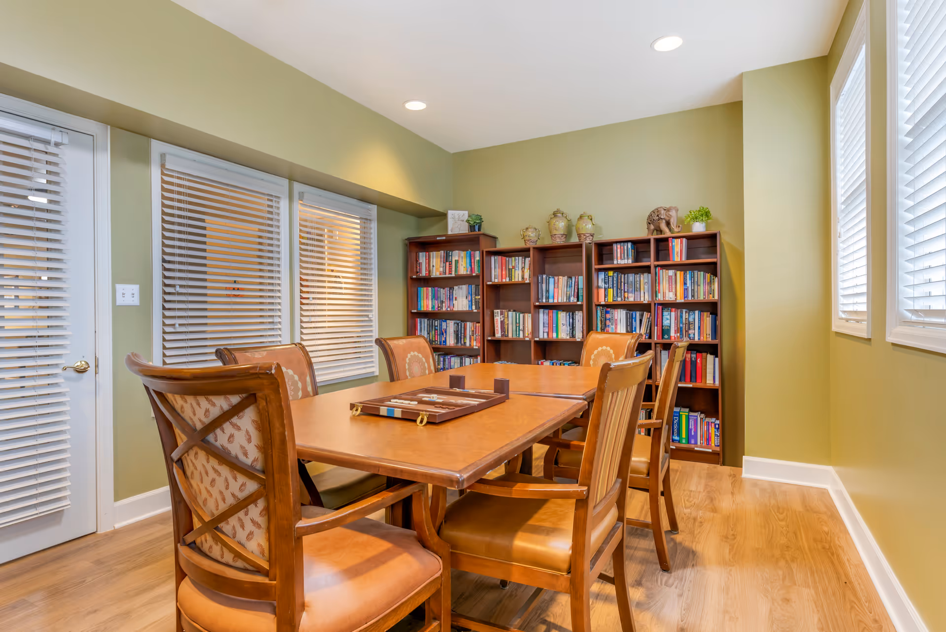 A wooden dining table with chairs in a room featuring bookshelves against a green wall and windows with blinds.