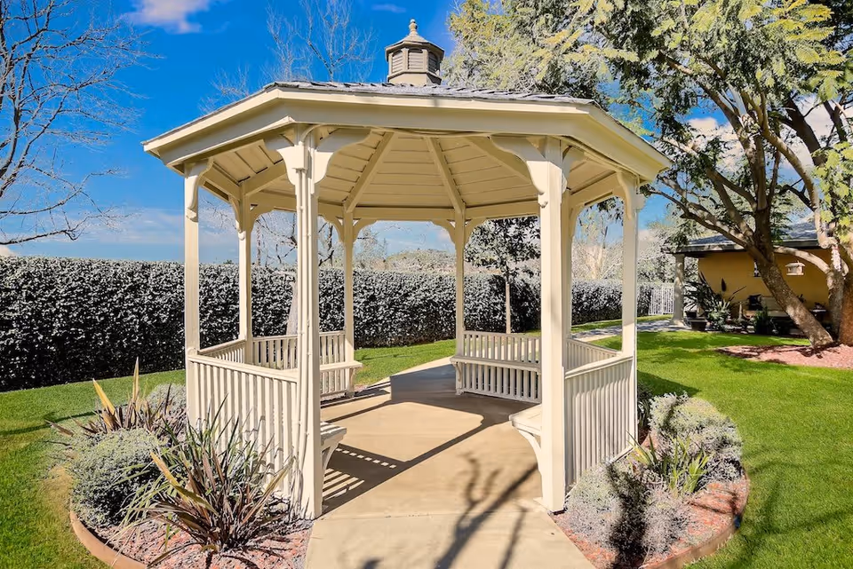 A white wooden gazebo with built-in benches situated on a concrete path surrounded by green grass, shrubs, and trees under a clear blue sky.