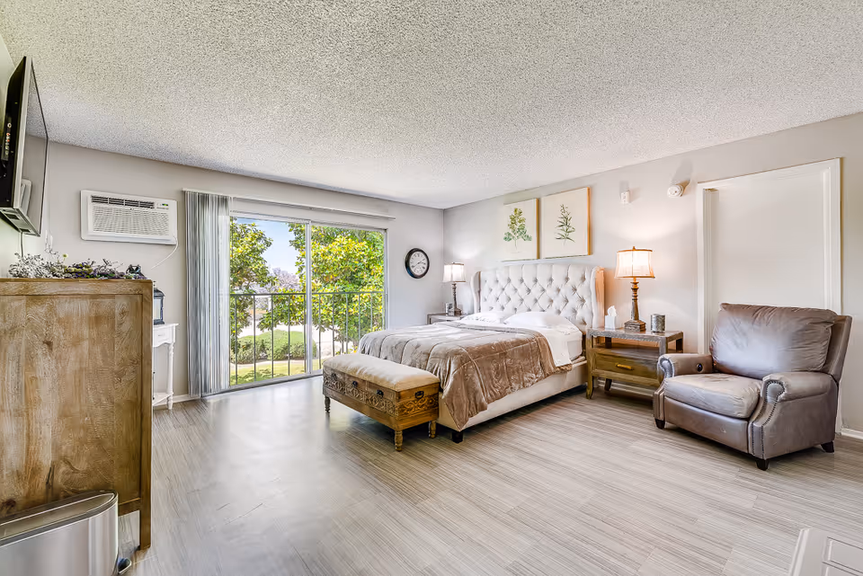 A bright and spacious bedroom with a large tufted bed covered with a beige blanket and white pillows. There is a wooden chest at the foot of the bed, two wooden nightstands with lamps on either side, a comfortable leather armchair, and a dresser with a TV mounted above it. A large sliding glass door with vertical blinds opens to a view of green trees outside. The room has light-colored walls and wood flooring.