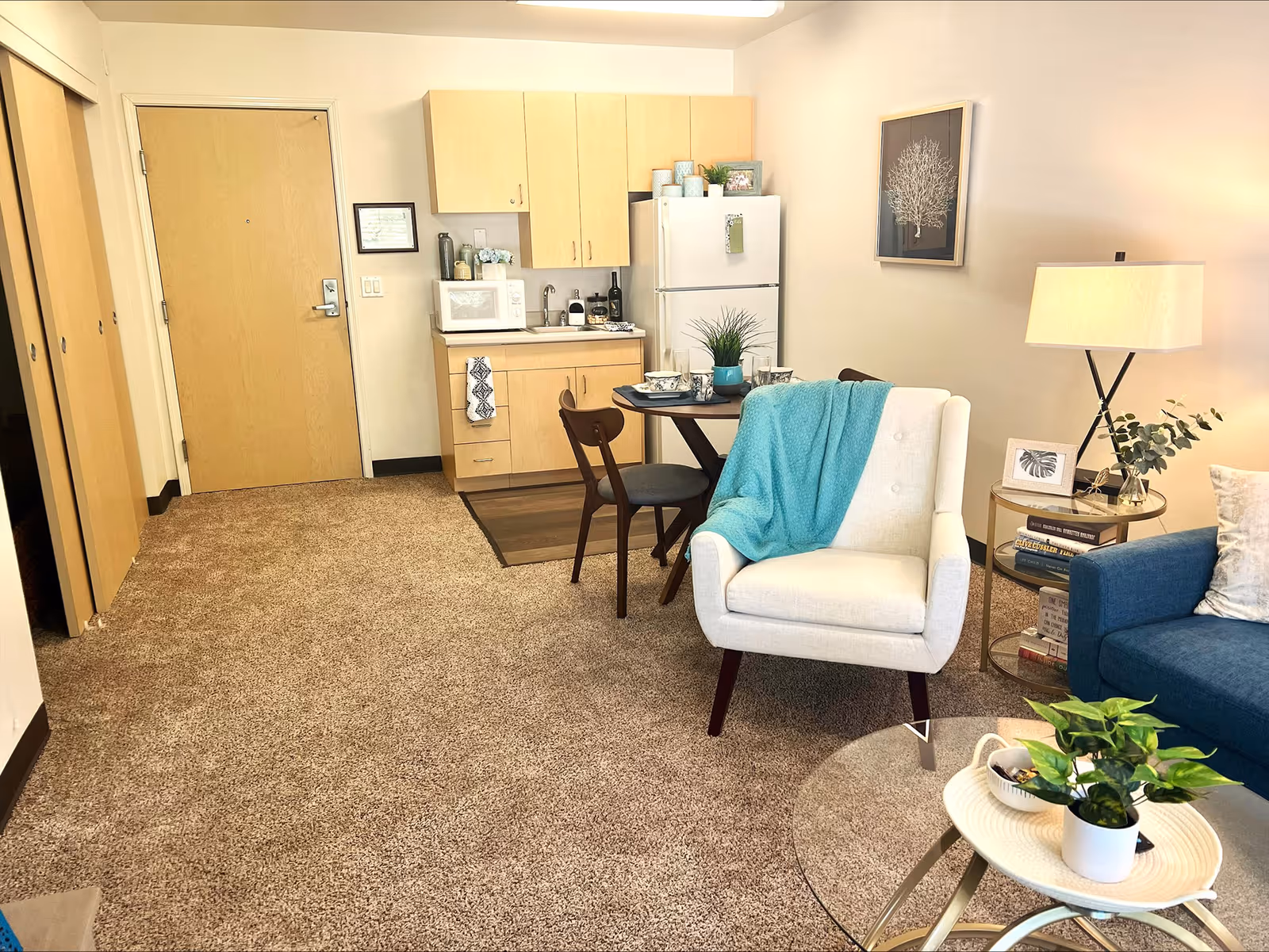 A cozy living space featuring a small kitchen area with light wood cabinets, a white refrigerator, and a microwave. In front of the kitchen is a round dining table set with dishes and a plant centerpiece, accompanied by two chairs. To the right, there is a white armchair draped with a turquoise throw blanket, a blue sofa, and a round glass coffee table with a small potted plant. A side table with a lamp, books, and framed pictures is next to the armchair. The room has beige carpet and light-colored walls with a framed artwork hanging above the armchair.