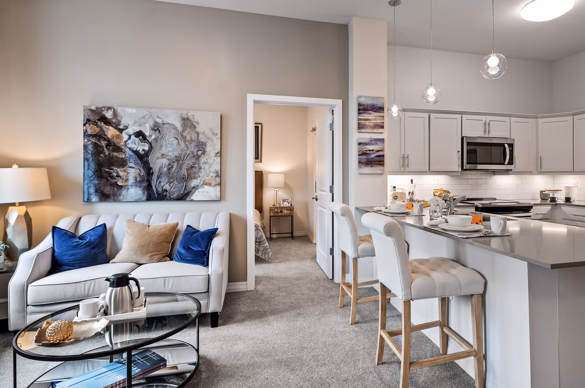 A modern living area with a beige sofa adorned with two blue and one brown throw pillows, a glass coffee table with a silver coffee pot and cup, and a lamp on a side table. The room opens into a bedroom with a bed and a nightstand with a lamp. To the right is a kitchen with white cabinets, a microwave, and a breakfast bar with two cushioned bar stools. The space is well-lit with pendant lights and neutral-colored walls.