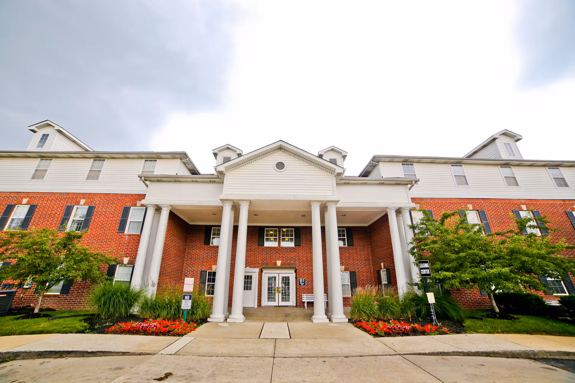 Front exterior view of a large brick building with white columns and a covered entrance. The building has multiple windows with black shutters and dormer windows on the roof. There are landscaped flower beds with red flowers and green bushes on either side of the entrance. The sky is cloudy.