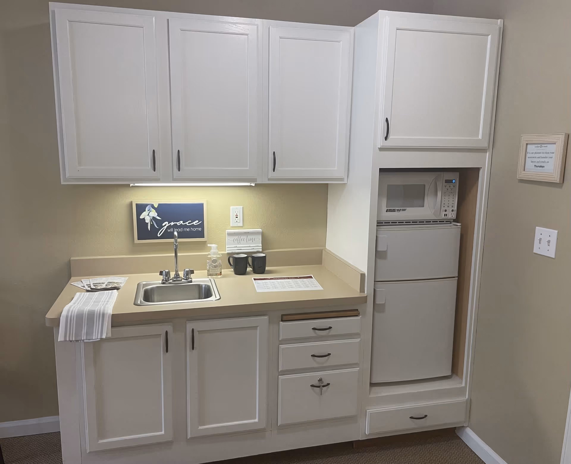 A small kitchenette area with white cabinets, a stainless steel sink, a microwave, and a mini refrigerator. There are two black mugs, a soap dispenser, and a striped towel on the counter. A sign above the sink reads 'grace will lead me home'.