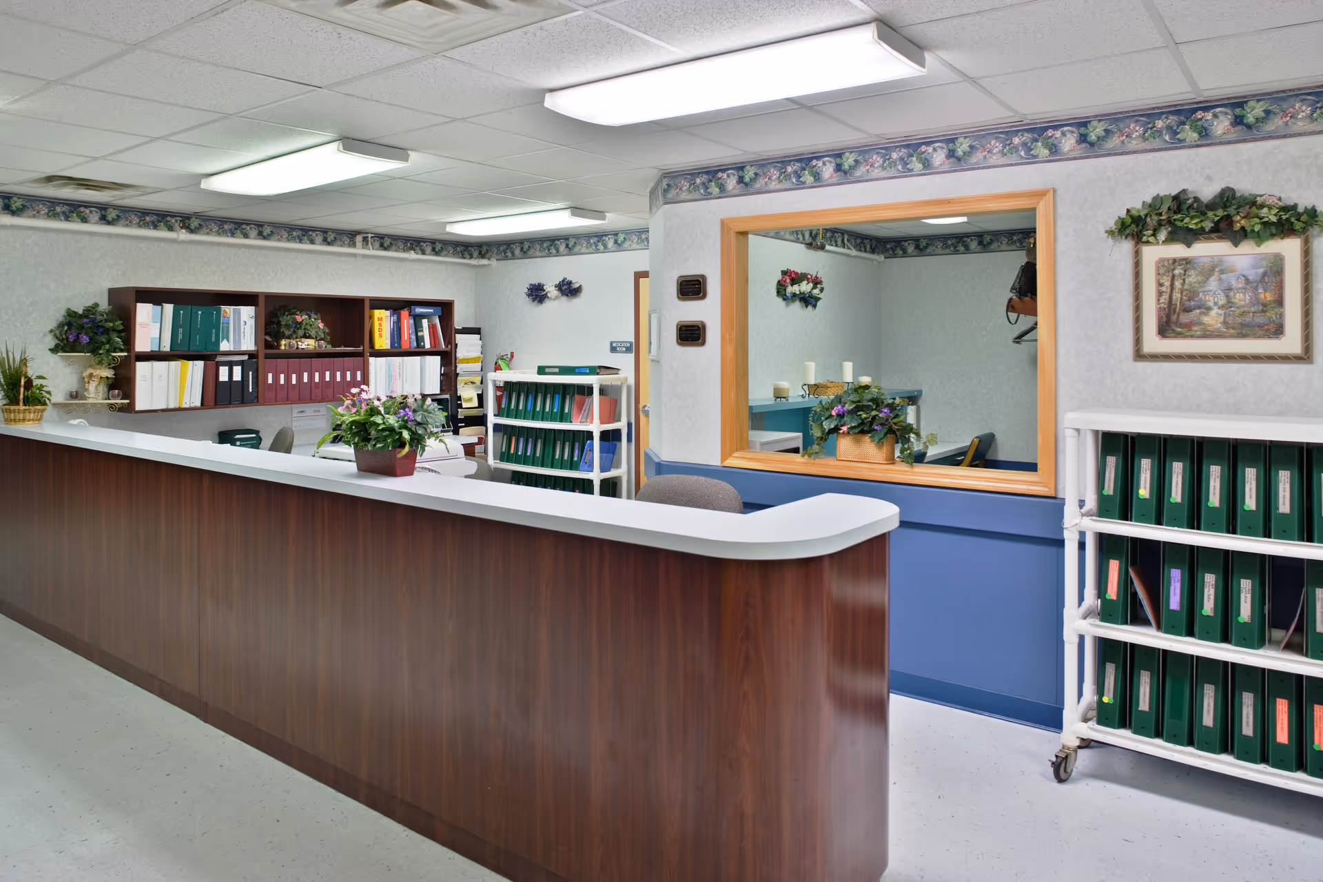 Reception desk and office area with shelving of binders, potted plants, and a service window.