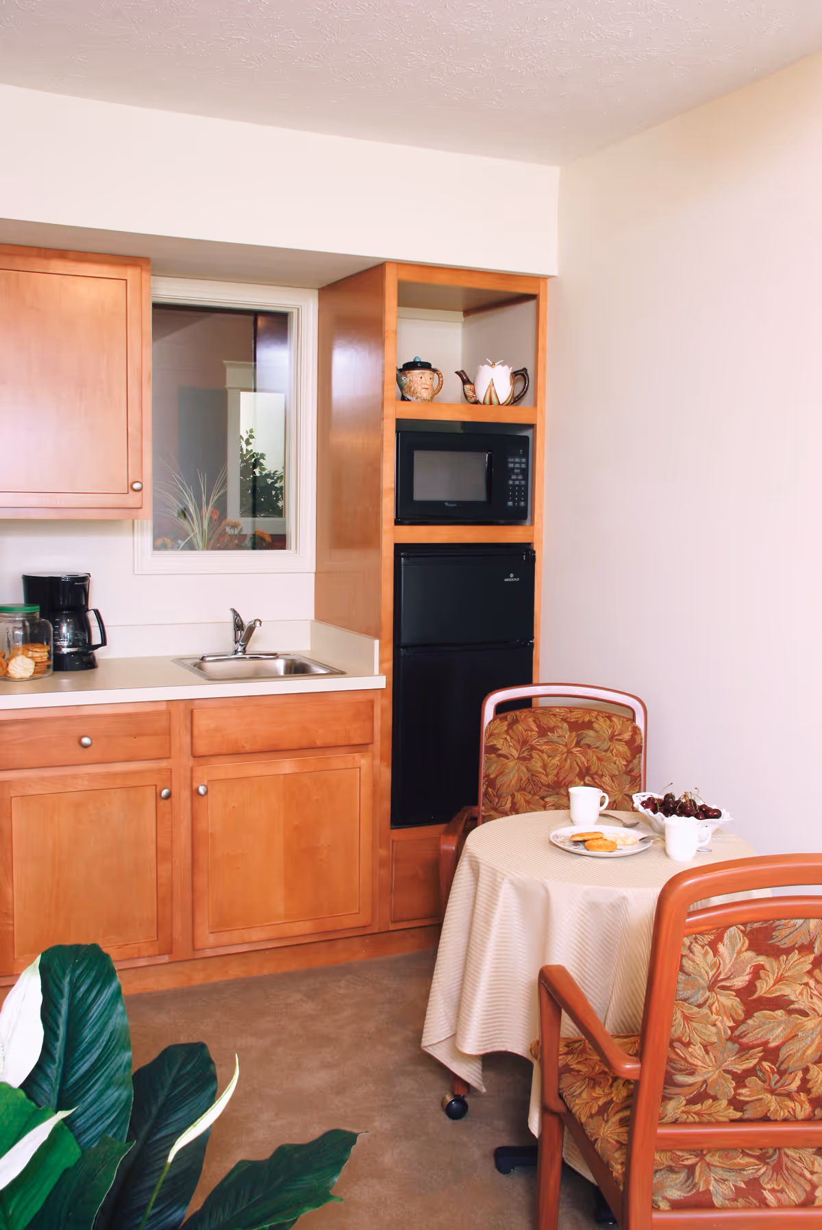 A small kitchenette area with wooden cabinets, a microwave, a refrigerator, and a dining table set for two.