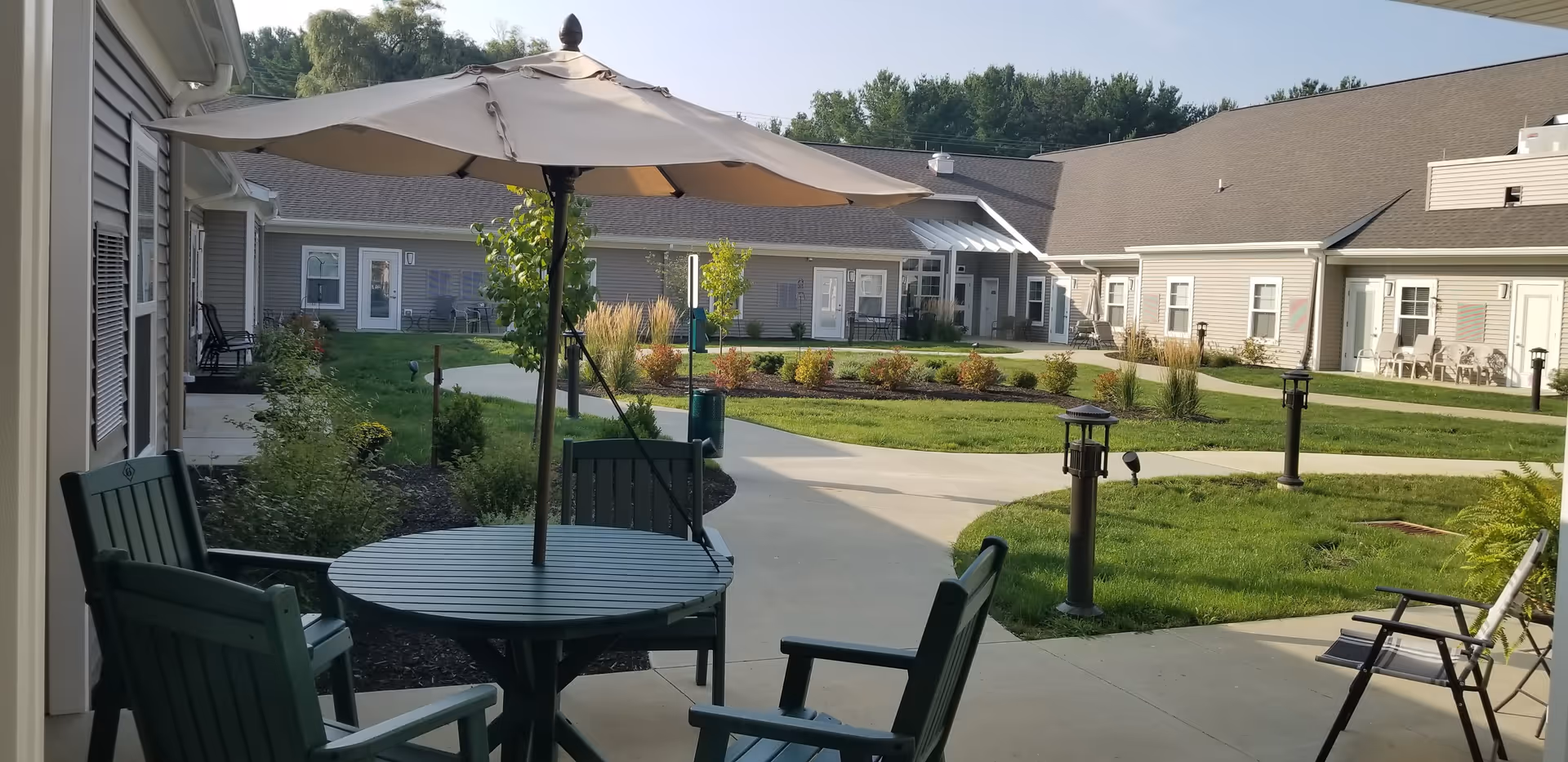 Outdoor courtyard area at The Gables of Green featuring a round table with four chairs and a large beige umbrella. The courtyard is surrounded by a single-story building with multiple doors and windows, landscaped with grass, shrubs, and small trees. Concrete walkways wind through the green space under a clear sky.