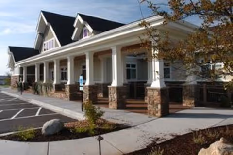 Front exterior of a senior living facility with a covered porch supported by columns, stone accents, parking spaces, and landscaped beds.