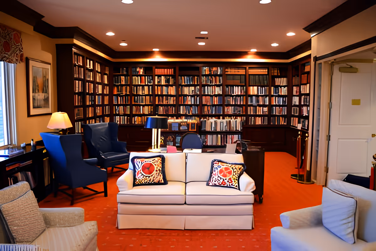 Cozy library-style common room with floor-to-ceiling bookshelves, a central white sofa with colorful pillows, armchairs and lamps.