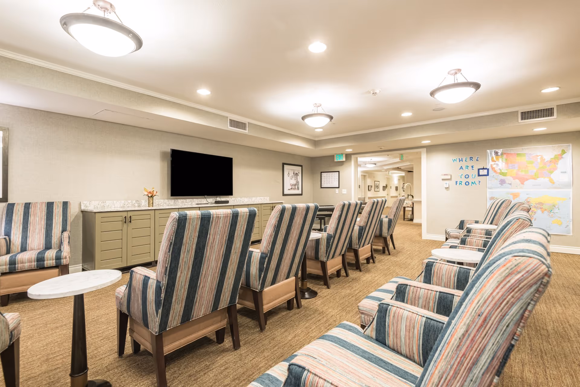 A well-lit common area with multiple striped upholstered armchairs arranged in rows facing a wall-mounted flat screen TV. There are small round tables between some chairs. The walls are light-colored with framed pictures and two maps on one side, along with a sign that reads 'WHERE ARE YOU FROM?'. The room has a carpeted floor and ceiling lights.