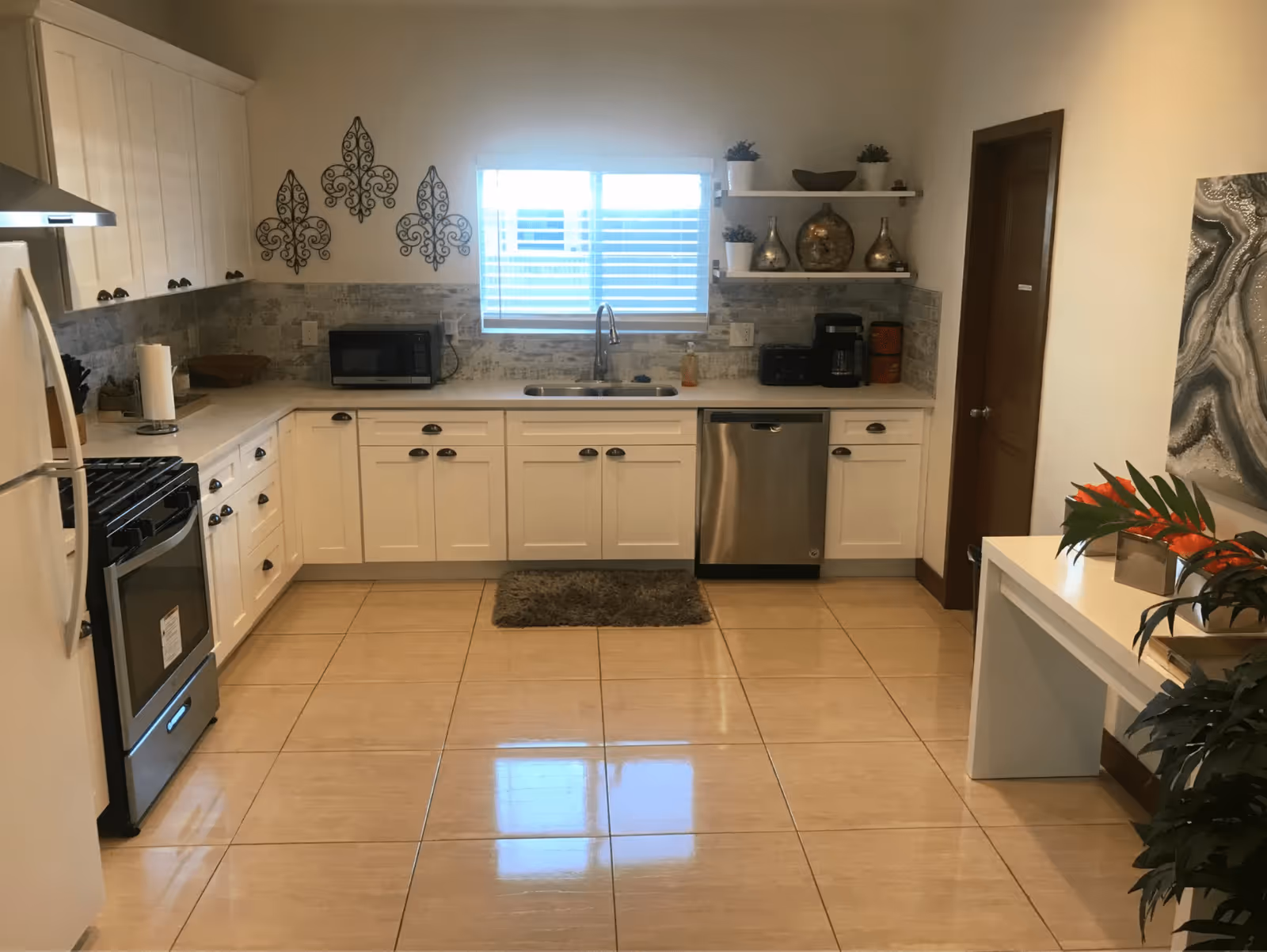 A clean and modern kitchen with white cabinets, a stainless steel stove and dishwasher, a microwave on the counter, a double sink under a window with blinds, decorative wall art, and shelves with plants and vases. There is a small rug in front of the sink and a white table with a plant and orange flowers on the right side.