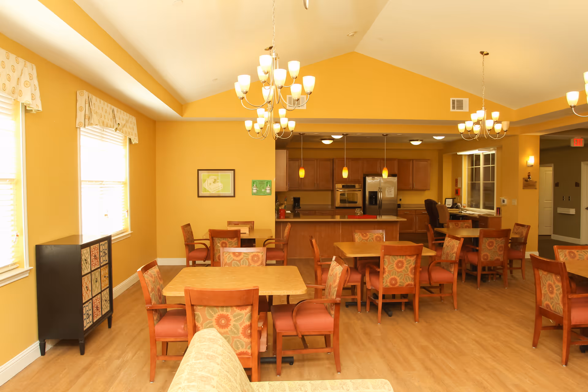 A bright and spacious dining area in an assisted living facility with multiple wooden tables and chairs featuring floral upholstery. The room has yellow walls, large windows with valances, and a kitchen area in the background equipped with stainless steel appliances and pendant lighting. Several chandeliers hang from the ceiling, illuminating the space.