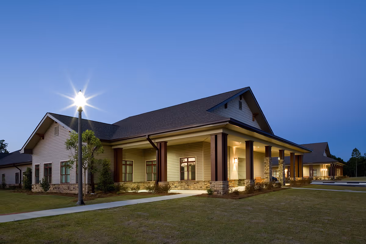 Exterior front of a single-story building at dusk with a covered, lit porch, a lamp post, and a grassy lawn.