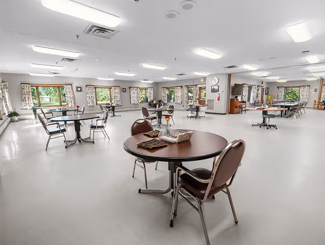 Large bright dining/activity room with round tables, chairs, and windows overlooking greenery.