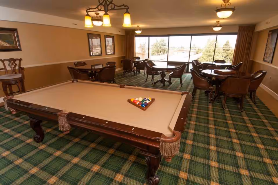 Carpeted common room with a pool table in the foreground and round tables and chairs by large windows.