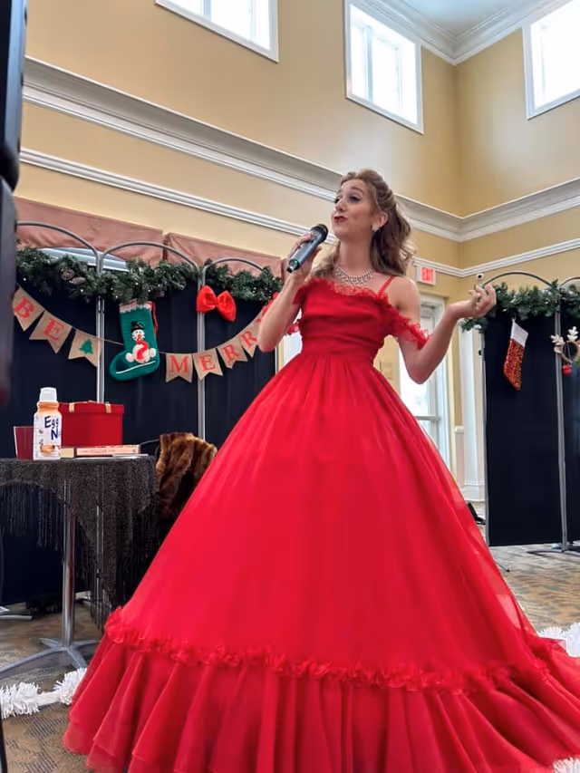 A woman in a bright red, elegant ball gown is singing into a microphone in a decorated indoor space. Behind her, there are festive Christmas decorations including a garland with stockings and a banner that reads 'BE MERRY'. The room has high ceilings with large windows letting in natural light.