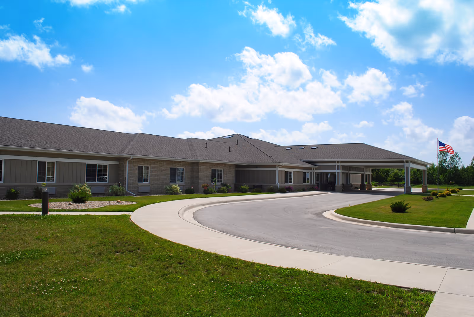 Exterior view of a single-story senior living facility building with a covered entrance driveway, surrounded by green grass and landscaping under a partly cloudy blue sky. An American flag is visible on a flagpole near the entrance.