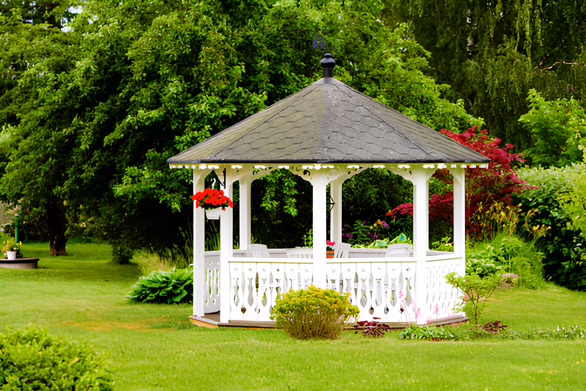 A white wooden gazebo with a shingled roof situated in a lush green garden surrounded by various trees, bushes, and colorful flowers. The gazebo has hanging red flowers and white chairs inside.