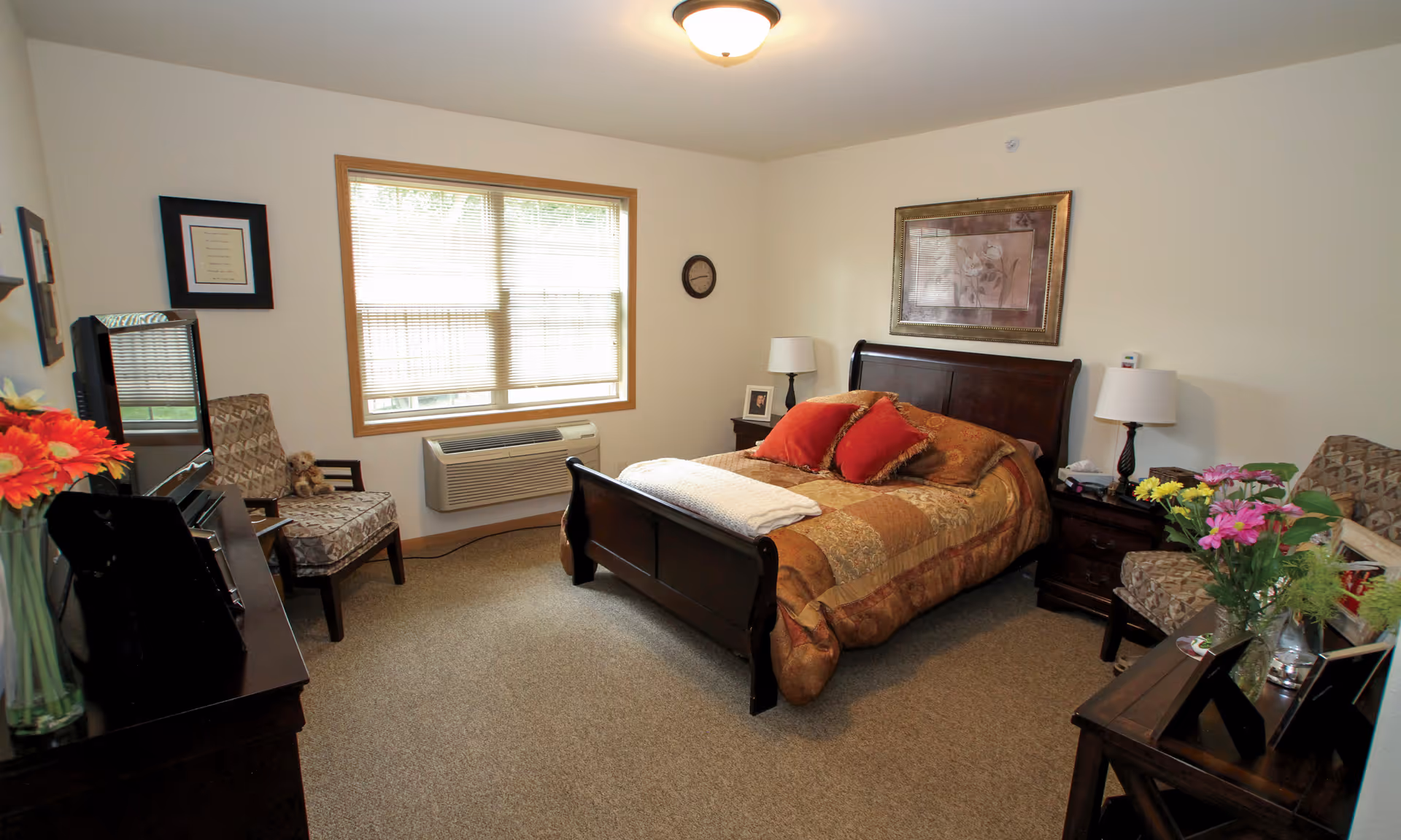 Sunlit furnished bedroom with a wooden bed, nightstands, chairs, dresser, and a window air conditioner.