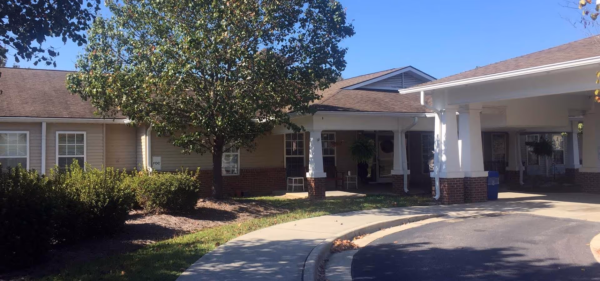 Exterior view of Hamlet House senior living facility showing a covered entrance with white columns and brick bases, surrounded by green bushes and a tree under a clear blue sky.