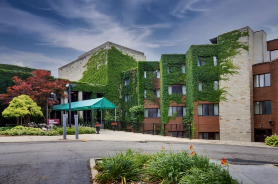 Exterior view of a multi-story building covered partially with green ivy. The building has a stone and wood facade with several windows. There is a green canopy over the entrance, surrounded by landscaped greenery and trees under a partly cloudy sky.