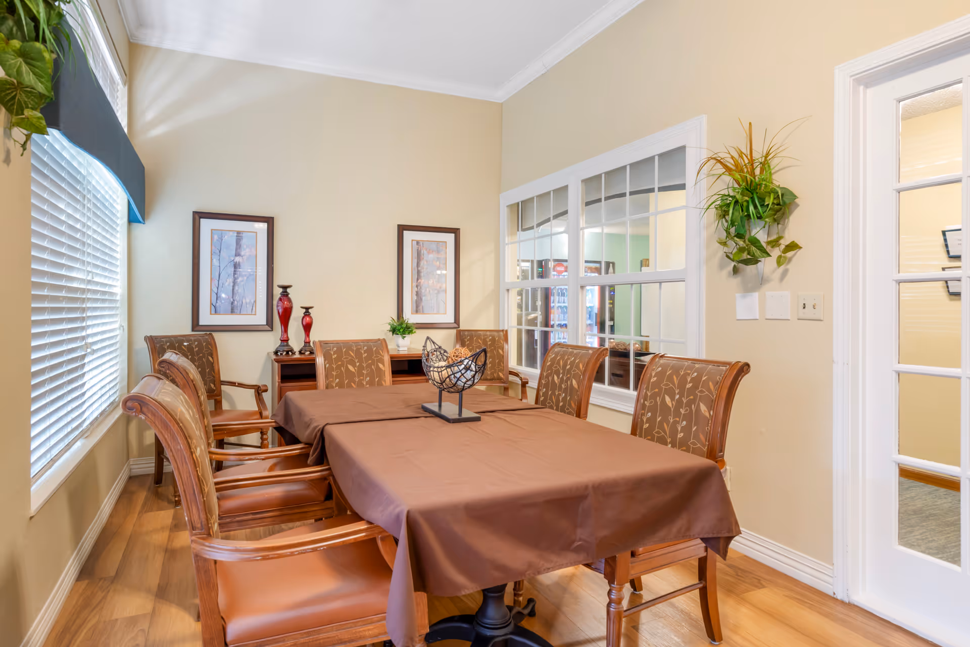 A dining room with a rectangular table covered by a brown tablecloth surrounded by six wooden chairs with patterned cushions. The room has light beige walls, two framed pictures on the far wall, a small side table with decorative items, a large window with blinds on the left, and a glass-paneled door on the right. A green plant is mounted on the wall near the door.