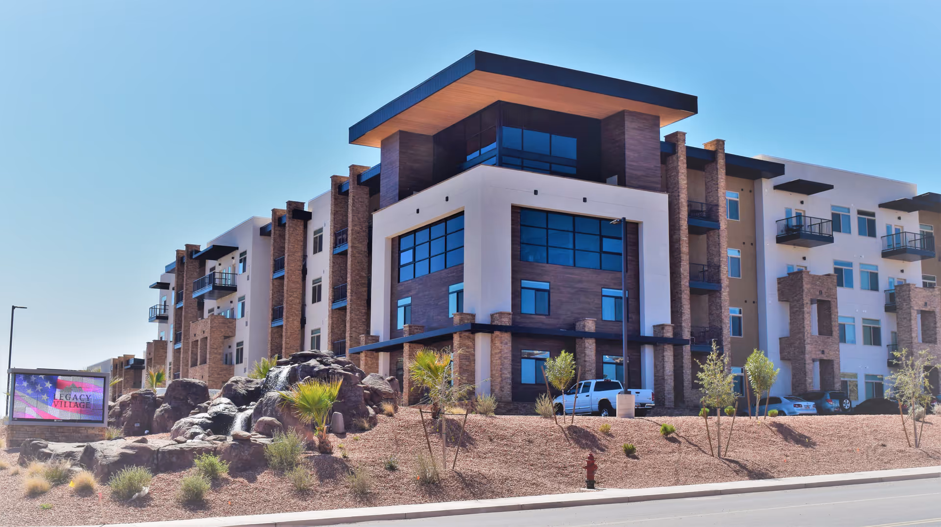 Exterior view of a modern multi-story senior living facility building with large windows, balconies, and a landscaped area with rocks and small plants in front. A sign near the road reads 'Legacy Village'.