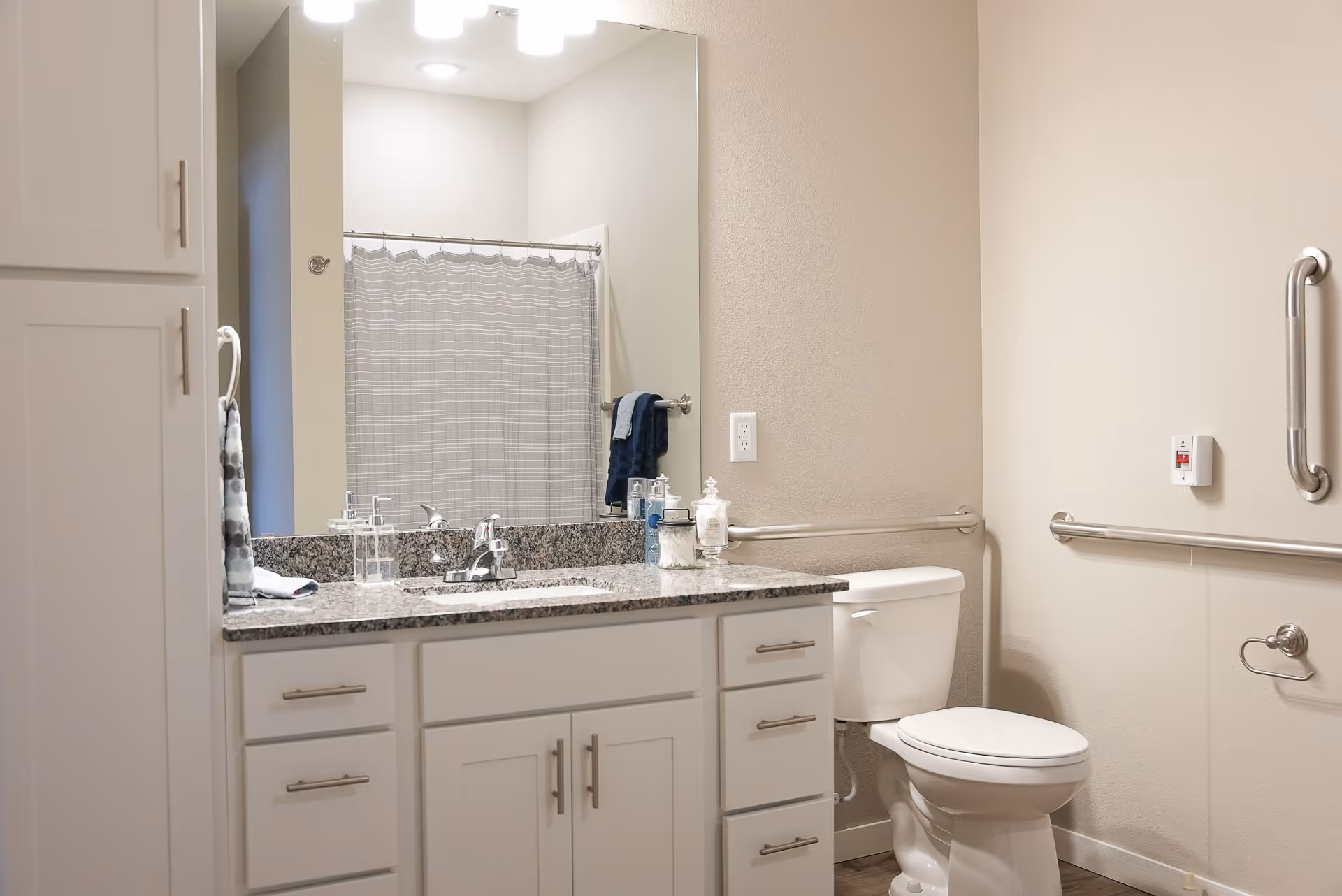 A clean and modern bathroom with a granite countertop vanity featuring a sink, soap dispenser, and decorative jars. There is a large mirror above the vanity, a toilet with grab bars on the walls for accessibility, a towel hanging on a ring, and a shower with a curtain in the background.