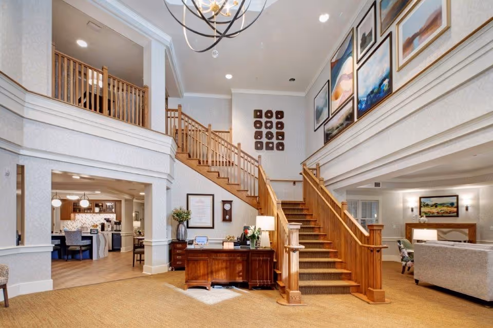 Interior view of a senior living facility lobby featuring a wooden staircase with carpeted steps leading to an upper floor. The area is decorated with framed artwork on the walls, a wooden desk with a lamp and flowers, and a seating area with couches and chairs. The space is well-lit with ceiling lights and has a warm, inviting atmosphere.
