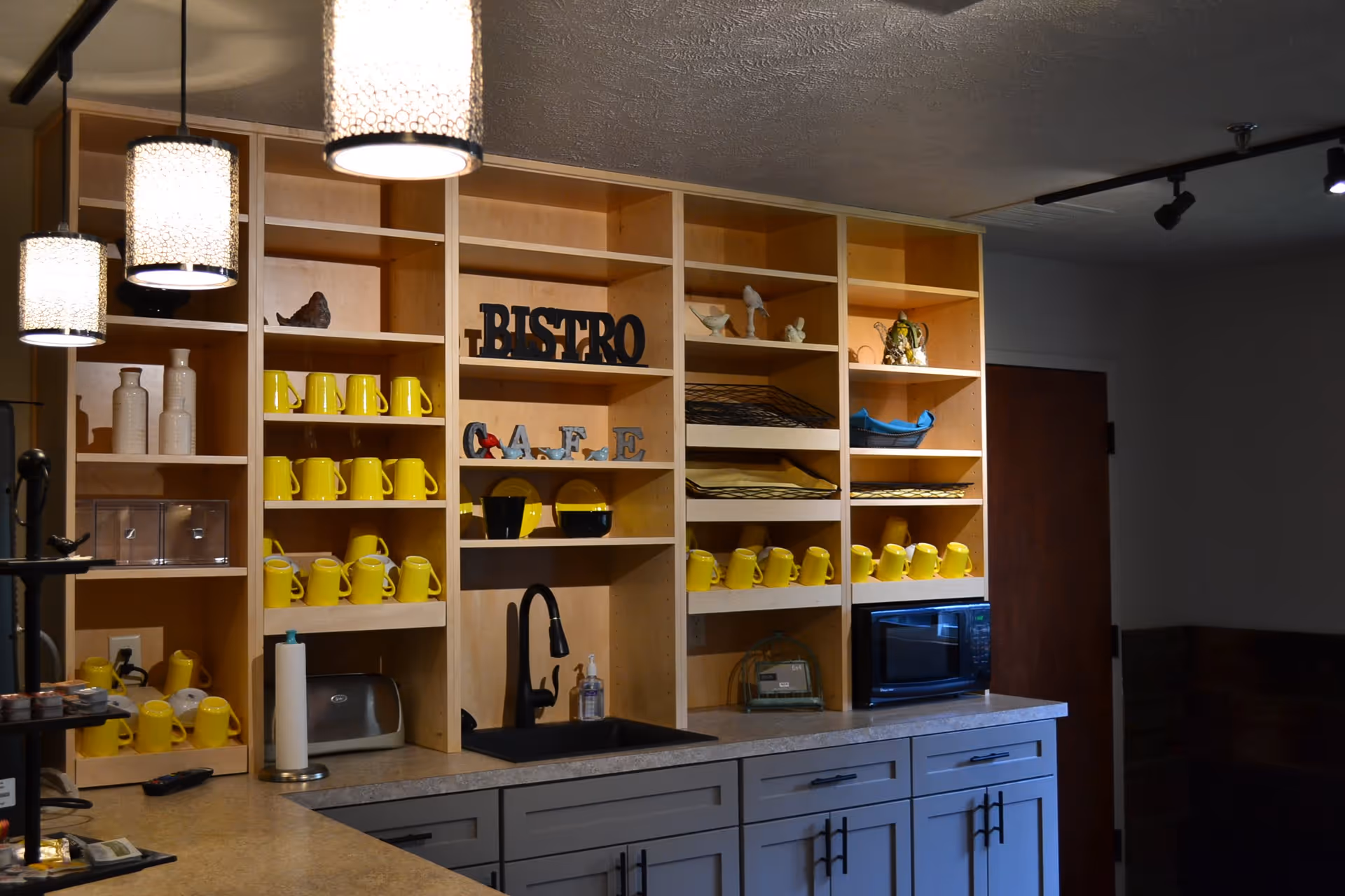 Interior view of a kitchen or café area with wooden shelves holding yellow mugs, decorative items, and signs that say BISTRO and CAFE. There is a countertop with a sink, a microwave, and hanging pendant lights above.