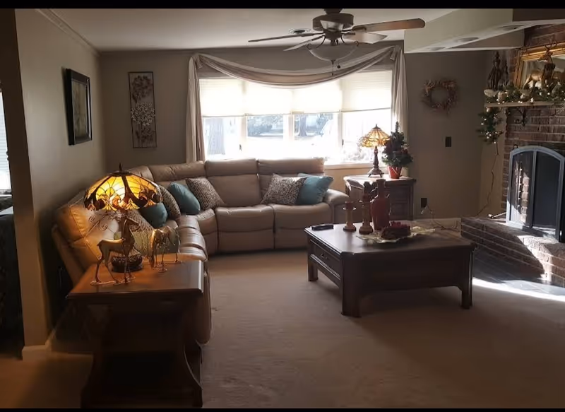 Cozy living room with a large beige sectional sofa adorned with decorative pillows, a wooden coffee table with ornaments, a side table with a stained glass lamp and horse figurines, a brick fireplace decorated with garland and a wreath, and a large window with curtains letting in natural light.