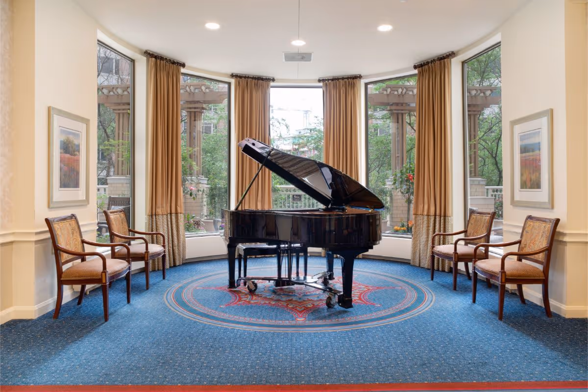 A bright room with a black grand piano placed on a decorative blue carpet with red and gold patterns. The room has large bay windows with beige curtains, allowing natural light to enter and showing an outdoor pergola and greenery. Four wooden chairs with upholstered seats and backs are arranged symmetrically, two on each side of the piano. The walls are light-colored and adorned with framed landscape paintings.