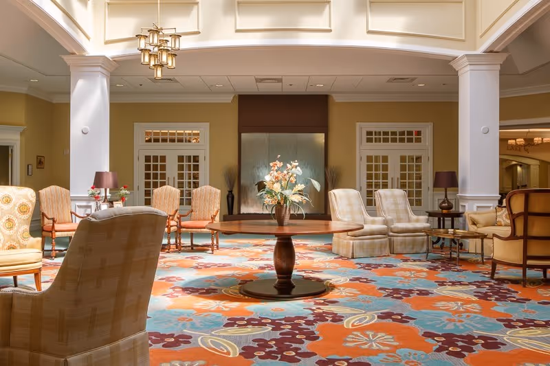 A spacious and well-lit common area with a colorful floral carpet, multiple upholstered chairs arranged in small groups, a round wooden table with a flower arrangement in the center, and large white columns supporting a high ceiling with a chandelier.