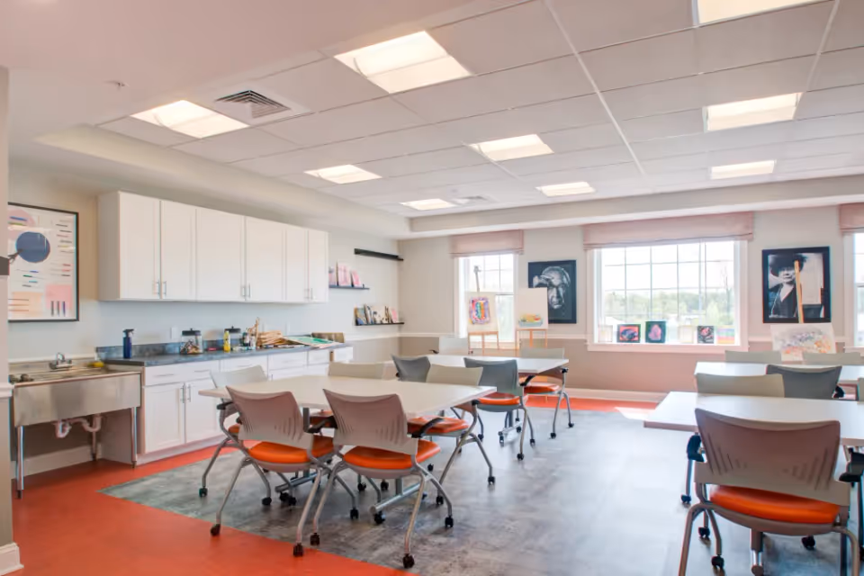 Communal activity room with tables and orange-seated chairs, a kitchenette with white cabinets, and large windows displaying artwork.