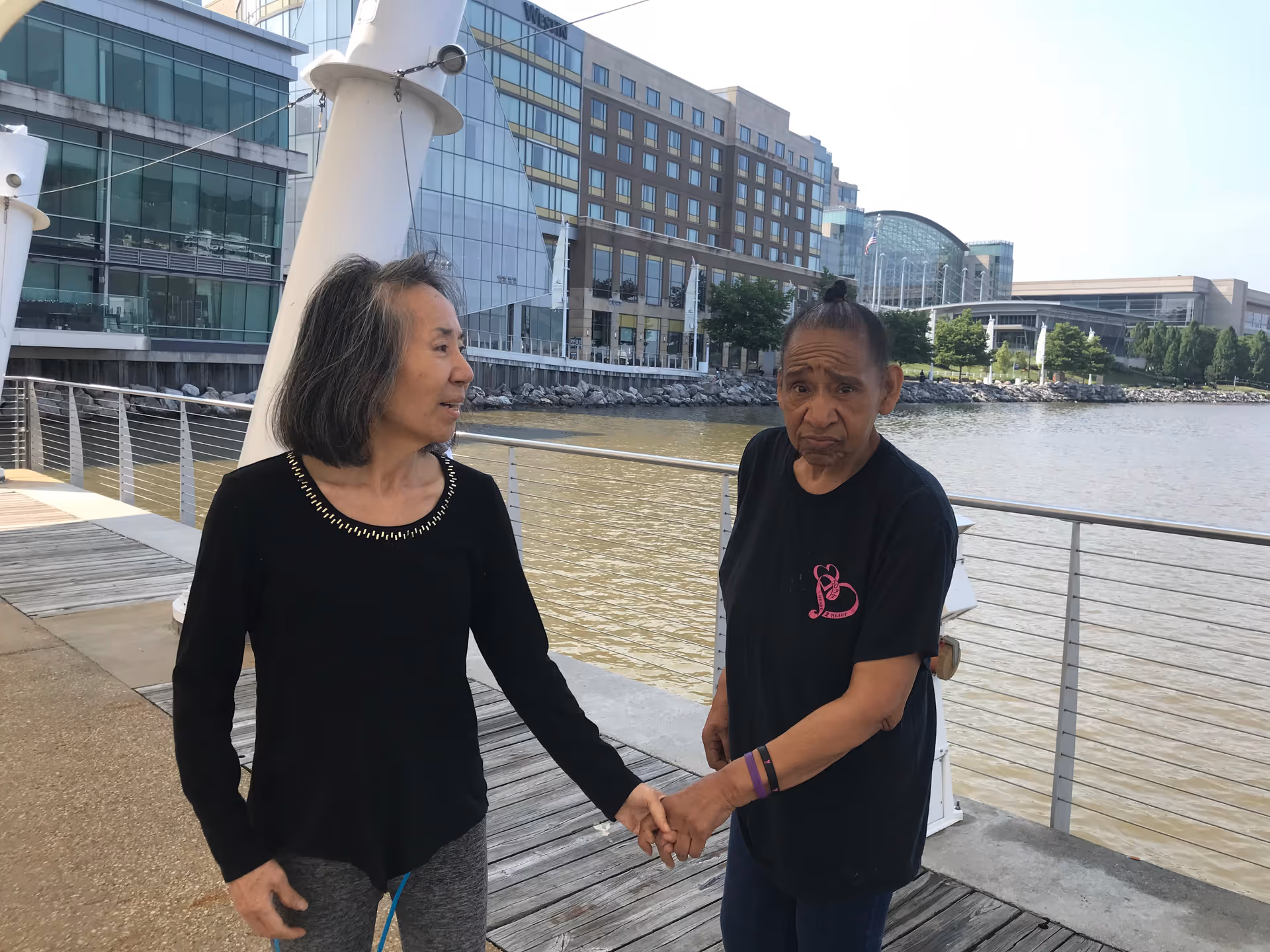 Two elderly individuals holding hands and standing on a wooden boardwalk by a waterfront with modern buildings in the background.