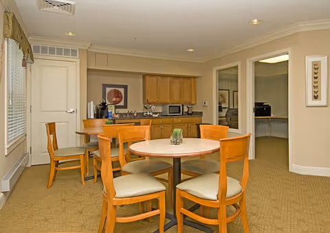 A cozy interior room with two round tables surrounded by wooden chairs with cushioned seats. In the background, there is a kitchenette area with wooden cabinets, a microwave, coffee maker, and a framed picture on the wall. Two doorways lead to adjacent rooms, one with a desk and printer visible. The room has beige walls, carpeted floor, and recessed ceiling lights.