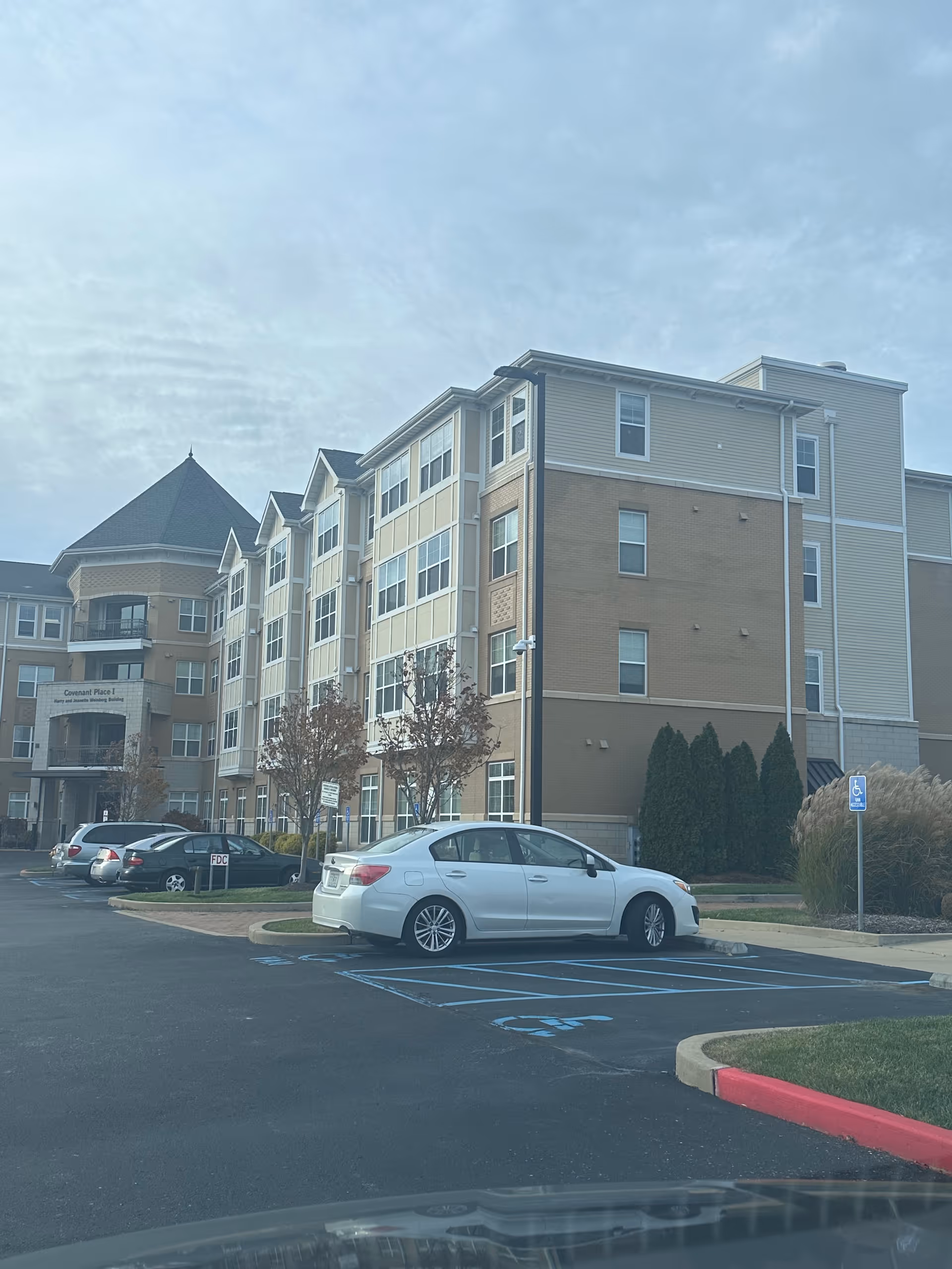 Front view of a multi-story senior living building with parked cars in the parking lot under a cloudy sky.