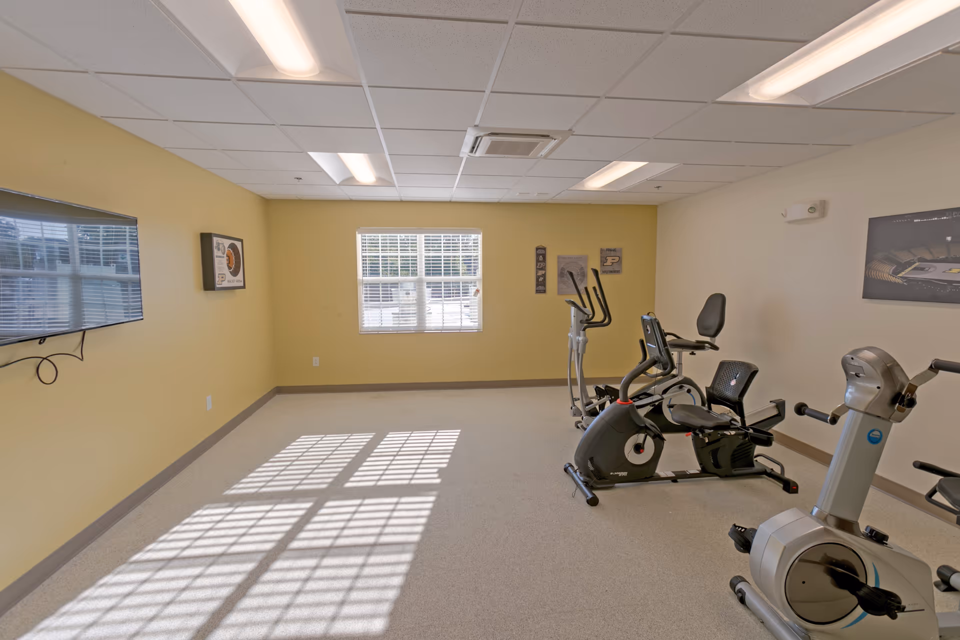 A small fitness room with exercise equipment including stationary bikes and an elliptical machine. The room has yellow and white walls, a window letting in natural light, a wall-mounted TV, and framed pictures on the walls.