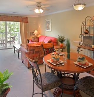 A cozy living and dining area with a round wooden dining table set for four, featuring plates, napkins, and glasses. The living room has a red sofa, patterned armchairs, a side table with a lamp, and a decorative metal shelf. Large sliding glass doors lead to a balcony with outdoor seating, allowing natural light to fill the room.