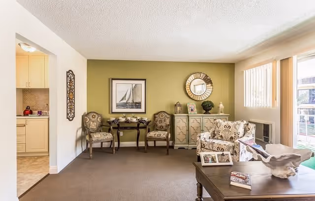 A cozy living room area with two patterned armchairs, a small wooden table with a decorative shell and photo frames, and a sideboard with a round mirror and decorative items. The room has a green accent wall with a framed sailboat picture, beige carpet, and vertical blinds covering a window and sliding glass door leading outside. A kitchen area is partially visible to the left.
