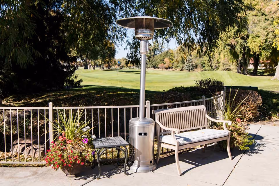 Outdoor patio area with a metal bench with a cushion, a tall outdoor heater, a small metal table, and potted plants with flowers. In the background, there is a green lawn and trees under a clear sky.