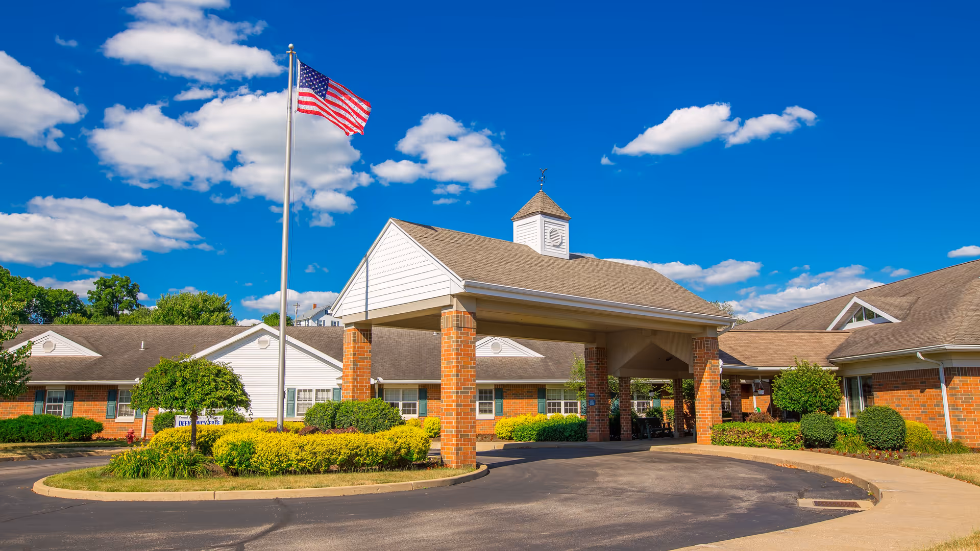 Front entrance of a brick nursing and rehabilitation center with a covered portico, American flag on a pole, and landscaped circular driveway.