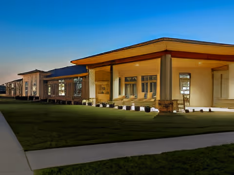 Exterior view of a single-story building with large windows and a covered porch area, surrounded by a well-maintained lawn and a paved walkway under a clear blue sky.