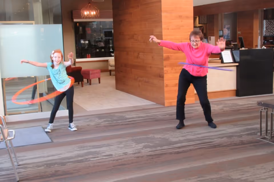 An elderly woman and a young girl are playing with hula hoops inside a modern indoor space with wooden and glass elements. The elderly woman is wearing a pink top and black pants, while the young girl is wearing a light blue shirt and black pants. The background shows a seating area with chairs and a reception desk.