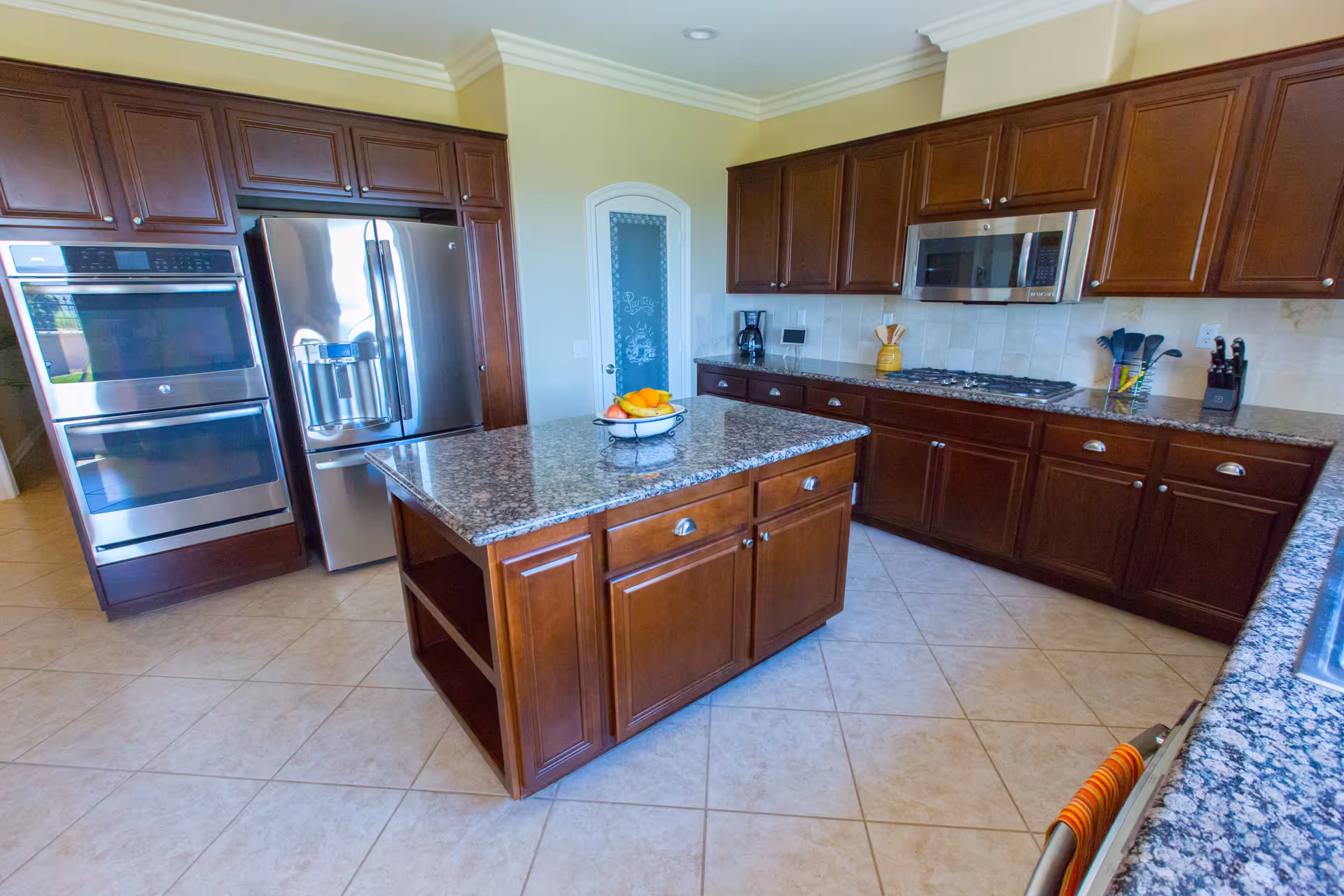 A spacious kitchen with dark wooden cabinets, a large granite island in the center with a bowl of fruit on top, stainless steel double ovens, a refrigerator, a microwave, a gas stove, and tiled flooring. There is a pantry door with frosted glass in the background.